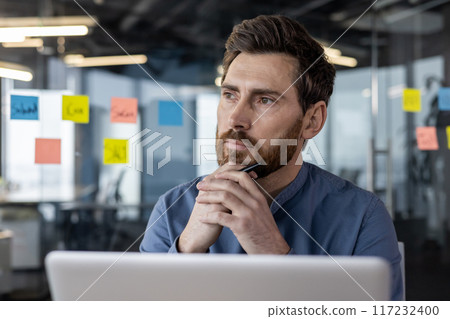 Businessman in modern office thinking while looking at laptop and holding pen. Professional reflecting on work. Business environment with colorful sticky notes on glass wall. 117232400
