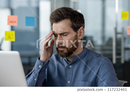 Frustrated businessman sitting in front of computer in modern office. Man holding head in pain feeling stressed and overwhelmed. Concept of work-related stress and mental health 117232401