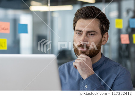Focused businessman with beard working on laptop in modern office space with glass walls and sticky notes. Emotion of concentration and problem-solving in professional environment 117232412