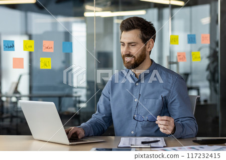Smiling businessman working on laptop in modern office environment. Man holding glasses and seated at desk with colorful sticky notes on glass wall in background. 117232415