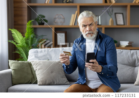 A senior Caucasian man looks puzzled while holding his credit card and smartphone, sitting on a sofa in a well-decorated living room. A senior Caucasian man looks puzzled while holding his credit card and smartphone, sitting on a sofa in a well-decorated living room. 117233131