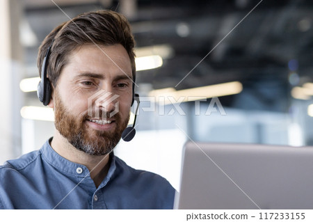 Confident businessman wearing headset while working on laptop in modern office. Engaged in video conference, virtual meeting, or customer support. Professional atmosphere 117233155