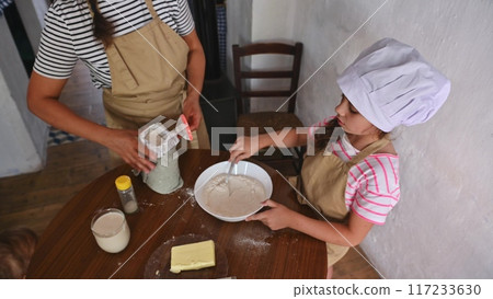 A mother and daughter spend quality time baking croissants together in a cozy kitchen. The little girl thoughtfully arranges baked goods while learning the art of cooking. A mother and daughter spend quality time baking croissants together in a cozy kitchen. The little girl thoughtfully arranges baked goods while learning the art of cooking. 117233630