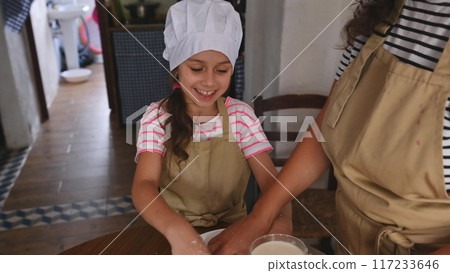 A mother and daughter spend quality time baking croissants together in a cozy kitchen. The little girl thoughtfully arranges baked goods while learning the art of cooking. 117233646