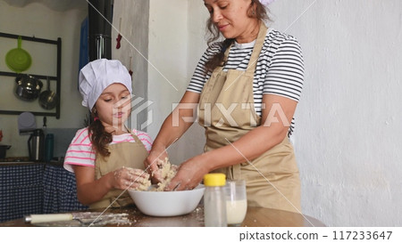 A mother and daughter spend quality time baking croissants together in a cozy kitchen. The little girl thoughtfully arranges baked goods while learning the art of cooking. 117233647