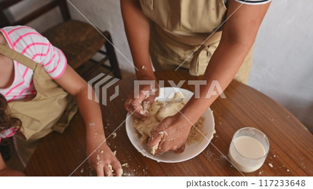 A mother and daughter spend quality time baking croissants together in a cozy kitchen. The little girl thoughtfully arranges baked goods while learning the art of cooking. 117233648