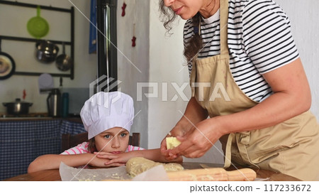 A mother and daughter spend quality time baking croissants together in a cozy kitchen. The little girl thoughtfully arranges baked goods while learning the art of cooking. 117233672