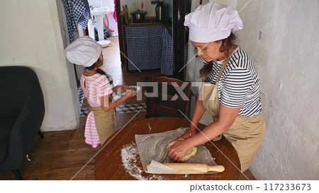 A mother and daughter spend quality time baking croissants together in a cozy kitchen. The little girl thoughtfully arranges baked goods while learning the art of cooking. 117233673