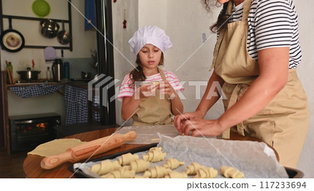 A mother and daughter spend quality time baking croissants together in a cozy kitchen. The little girl thoughtfully arranges baked goods while learning the art of cooking. 117233694