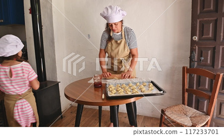 A mother and daughter spend quality time baking croissants together in a cozy kitchen. The little girl thoughtfully arranges baked goods while learning the art of cooking. 117233701