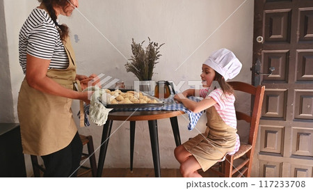 A mother and daughter spend quality time baking croissants together in a cozy kitchen. The little girl thoughtfully arranges baked goods while learning the art of cooking. A mother and daughter spend quality time baking croissants together in a cozy kitchen. The little girl thoughtfully arranges baked goods while learning the art of cooking. 117233708