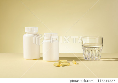 The photography at light yellow background features white unbranded supplement jars, a glass of water and yellow vitamin capsules. Front angle shot, blank space for adding text The photography at light yellow background features white unbranded supplement jars, a glass of water and yellow vitamin capsules. Front angle shot, blank space for adding text 117235827