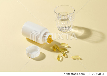 High angle shot at the yellow countertop with an unlabeled supplement bottle flat lay by a glass of water and plenty yellow capsules scattered. Mockup for advertisement, empty space to design High angle shot at the yellow countertop with an unlabeled supplement bottle flat lay by a glass of water and plenty yellow capsules scattered. Mockup for advertisement, empty space to design 117235831