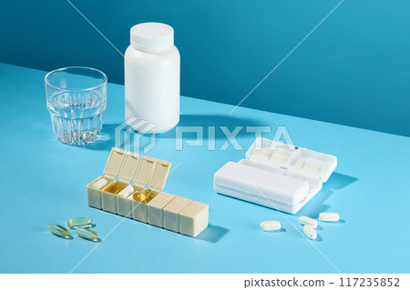 Baby blue table top contains several items prepared to drink medicine like daily pill box, vitamin capsule, mineral tablets, unlabeled pill jar and a glass of water. Photo for advertising, empty space 117235852
