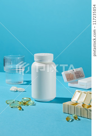 White plastic medicine bottle against on blue background, decorated around with glass of water and pill boxes containing vitamin of various colors and shapes by day for memory. Front view, copy space 117235854