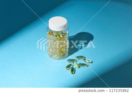 The transparent supplement bottle isolated on blue background, next to some flat lay vitamin supplement capsule. The photo was taken from above view with blank space for promotional The transparent supplement bottle isolated on blue background, next to some flat lay vitamin supplement capsule. The photo was taken from above view with blank space for promotional 117235862