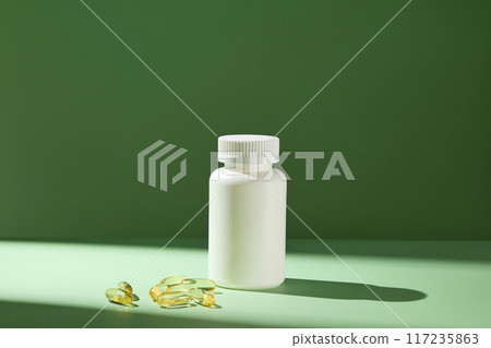Frontal photo taken on a green background with a streak of sun shining through an unlabeled white medicine bottle displayed in center, next to some flat lay supplement capsules. Advertising space 117235863