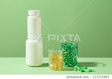 Two different sizes white medicine bottles placed on top of each other, glass beaker contains yellow and green vitamin capsules. Creative advertising mockup, front view and space for text 117235867