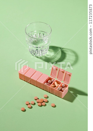 Illustration photo of pharmaceutical advertisement with a pink pill organizer placed next to a water glass on green table, contains pills and capsules. High angle shot, product presentation space Illustration photo of pharmaceutical advertisement with a pink pill organizer placed next to a water glass on green table, contains pills and capsules. High angle shot, product presentation space 117235873