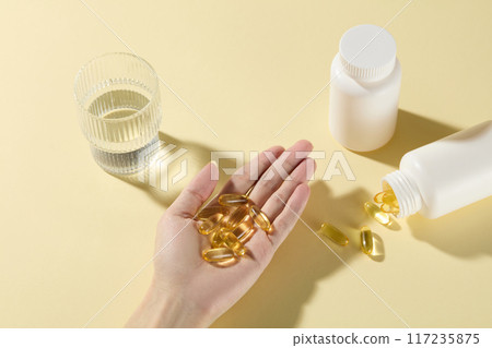 Medicine drinking preparation scene with a female hand holding yellow supplement capsules in palm, glass of water and unbranded white medicine bottles placed around. Top view, copy space for text Medicine drinking preparation scene with a female hand holding yellow supplement capsules in palm, glass of water and unbranded white medicine bottles placed around. Top view, copy space for text 117235875