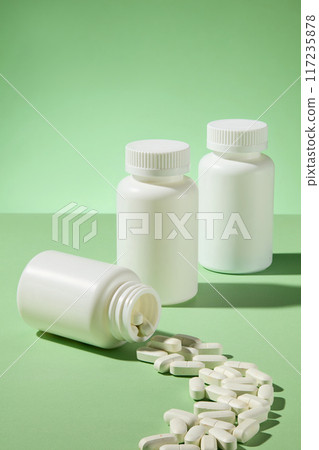White tablets from white unlabeled drug jar spilled as a line on the green table, in back of it is two mockup medicine bottles standing by side. Front angle shot, promotional space 117235878