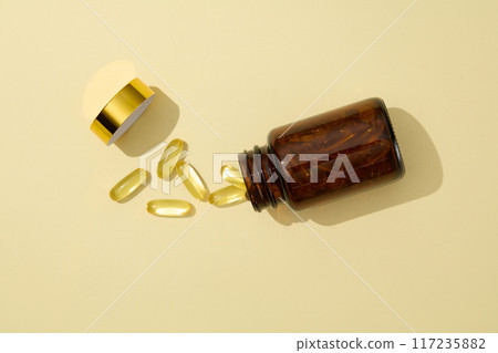 Flat lay amber medicine jar is opened and isolated on light yellow background, with some yellow soft fish oil capsule and golden lid scattered lay beside. Top view, blank space for text Flat lay amber medicine jar is opened and isolated on light yellow background, with some yellow soft fish oil capsule and golden lid scattered lay beside. Top view, blank space for text 117235882