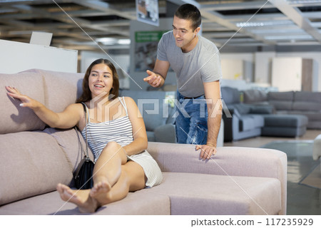 Happy woman sitting on couch in store while seller talking about model Happy woman sitting on couch in store while seller talking about model 117235929