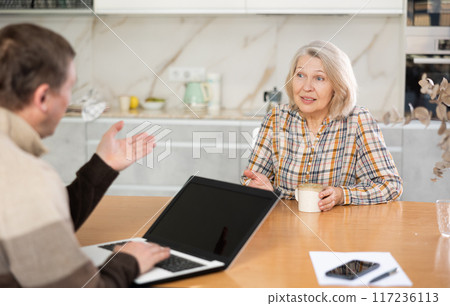 Male insurance agent tells an elderly woman the terms of an insurance contract. Offers to sign documents in room Male insurance agent tells an elderly woman the terms of an insurance contract. Offers to sign documents in room 117236113