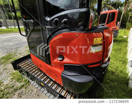 Red large excavators are stationed in a green field in front of a building Red large excavators are stationed in a green field in front of a building 117236695