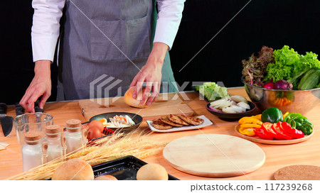Young Chef holding bread bun on cutting board with fresh vegetables, eggs, and spices on kitchen counter 117239268