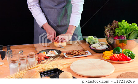Young Chef slicing bread bun on cutting board with fresh vegetables, eggs, and spices on kitchen counter Young Chef slicing bread bun on cutting board with fresh vegetables, eggs, and spices on kitchen counter 117239269