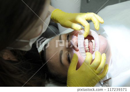 Dentist Closeup woman patient. Carious tooth treatment at dental clinic office. Nurse, doctor hand. Dentist making teeth cleaning, treating and canal filling. assistant spaying mouth 117239371