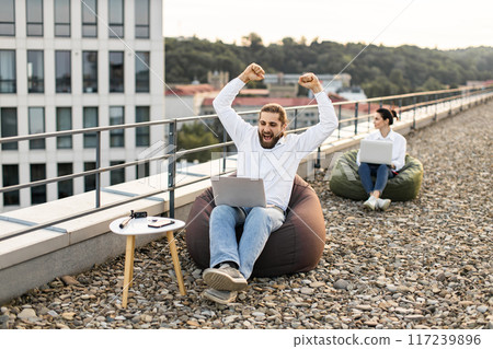 Excited man working on laptop outdoors on rooftop with colleague 117239896