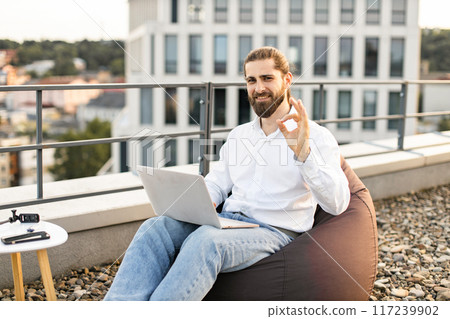 Smiling man working remotely on rooftop terrace with laptop Smiling man working remotely on rooftop terrace with laptop 117239902