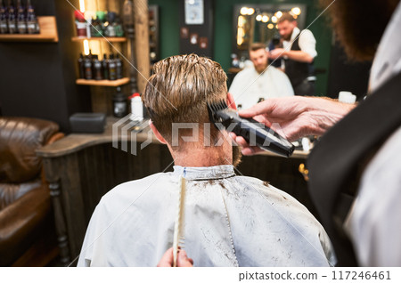 Professional barber using comb and electric shaver to cut side hair. Close-up view of male customer getting stylish haircut. Professional barber using comb and electric shaver to cut side hair. Close-up view of male customer getting stylish haircut. 117246461
