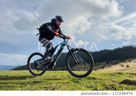 Cyclist man riding electric mountain bike outdoors. Male tourist biking along grassy trail in the mountains, wearing helmet and backpack. Concept of sport, active leisure and nature. 117246474