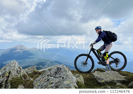 Cyclist man riding electric mountain bike outdoors. Male tourist biking along grassy trail in the mountains, wearing helmet and backpack. Concept of sport, active leisure and nature. 117246476