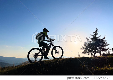 Cyclist man riding electric mountain bike outdoors. Male tourist biking along grassy trail in the mountains, wearing helmet and backpack. Concept of sport, active leisure and nature. 117246478