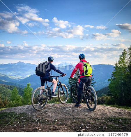 Two cyclists men riding electric bikes outdoors. Back view of male tourists resting on the top of hill, enjoying beautiful mountain landscape, wearing helmet and backpack. Concept of active leisure. 117246479