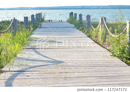 Wooden paved bridge to lake on beach. Sunny day and relaxation. Nature and sky. Background with perspective for design.  117247172
