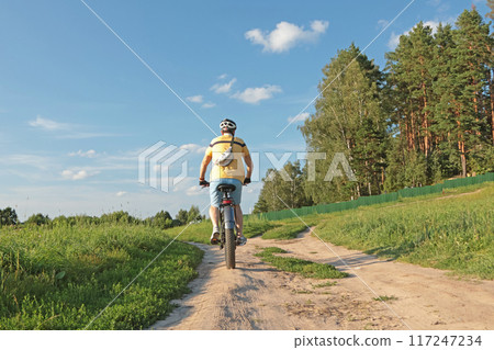 Cyclist in helmet with equipment on electric bicycle rides along path in field in countryside. Sports and health. Summer hobby. Cyclist in helmet with equipment on electric bicycle rides along path in field in countryside. Sports and health. Summer hobby. 117247234