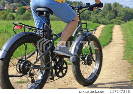 Cyclist on electric bicycle riding along path in field in countryside at sunset close-up. Sport and health. Summer hobby. Cyclist on electric bicycle riding along path in field in countryside at sunset close-up. Sport and health. Summer hobby. 117247236