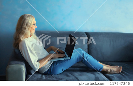 Modern caucasian mature woman working with laptop sitting on a couch in living room at home Modern caucasian mature woman working with laptop sitting on a couch in living room at home 117247246