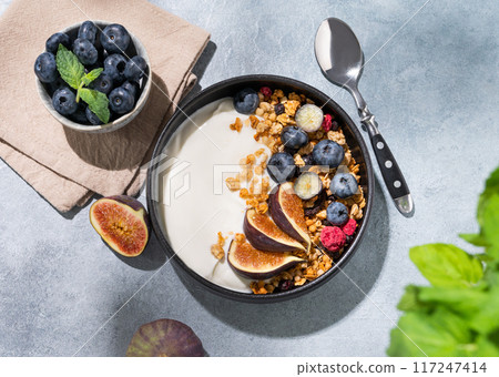 Natural yogurt with granola, berries and figs in a black bowl on a blue background with mint Natural yogurt with granola, berries and figs in a black bowl on a blue background with mint 117247414