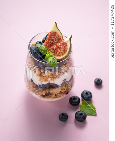 Natural yogurt with granola, figs and blueberries in a glass on a pink background. Natural yogurt with granola, figs and blueberries in a glass on a pink background. 117247428