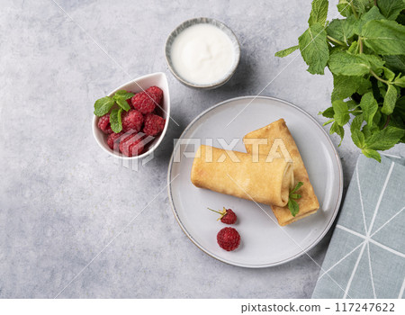 Homemade crepes with raspberry and sour cream on a blue plate on a light background with fresh mint. 117247622
