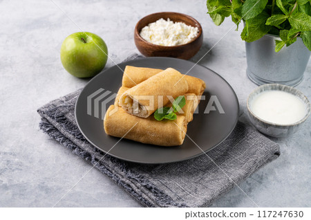 Homemade crepes roll with cottage cheese and apple on a plate on a blue background with sour cream Homemade crepes roll with cottage cheese and apple on a plate on a blue background with sour cream 117247630
