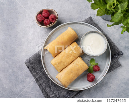 Homemade crepes with raspberry and sour cream on a plate on a light background with fresh mint  117247635