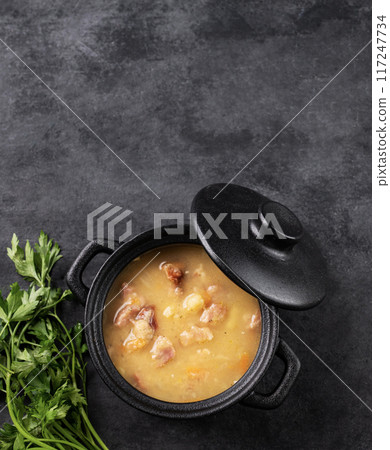 Pea soup with smoked ham in black soucepan on dark background with herb. 117247734