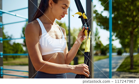 Confident, athlete woman holds onto TRX strap, basking in warmth of sun after intense outdoor training session. Confident, athlete woman holds onto TRX strap, basking in warmth of sun after intense outdoor training session. 117247967